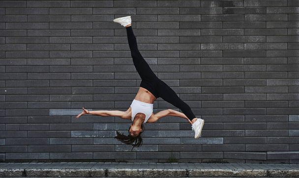 Doing crazy parkour and athletic stunts. Young sportive brunette with slim body shape against brick wall in the city at daytime. - Фото, изображение