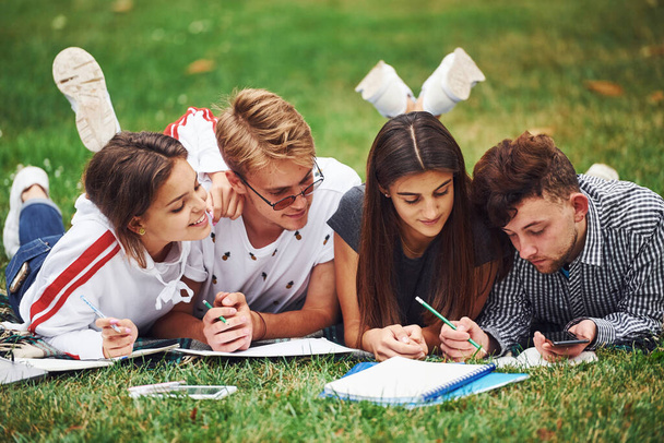 Writing on paper. front view. Group of young students in casual clothes on green grass at daytime. - Foto, imagen