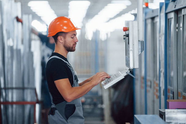 Using machine to program operations. Industrial worker indoors in factory. Young technician with orange hard hat. - Foto, Imagen