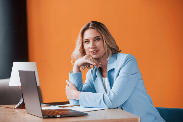 If you need advice, I can help. Businesswoman with curly blonde hair indoors in room with orange colored wall and wooden table. - Foto, imagen