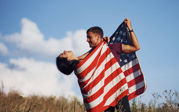 Αισθάνεται ελευθερία. Όμορφο ζευγάρι με αμερικανική σημαία έχουν μια καλή στιγμή σε εξωτερικούς χώρους στο πεδίο. - Φωτογραφία, εικόνα