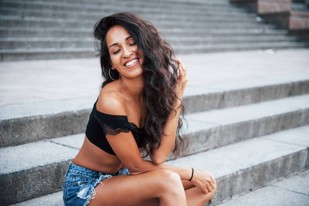 Toothy smile. Beautiful woman with curly black hair have good time in the city at daytime. - Zdjęcie, obraz