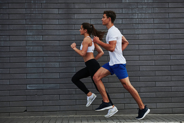 Side view. Man and woman have fitness day and running in the city at daytime near brick wall. - Foto, Imagen