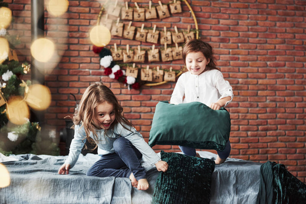 Look straight into the camera while holding the pillow. Little girls having fun on the bed with holiday interior at the background. - Foto, Imagen