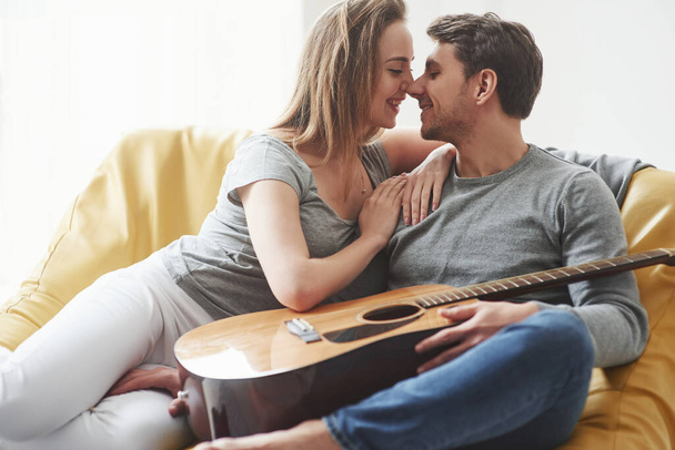 Giving a kiss. Guitar on the legs. Happy couple relaxing on the yellow sofa in the living room of their new house. - Фото, изображение