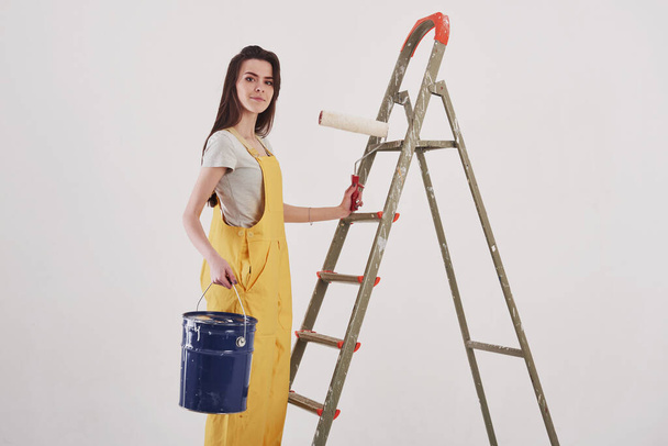 Beautiful brunette in yellow uniform and blue colored bucket stands near the ladder. - Foto, Imagem