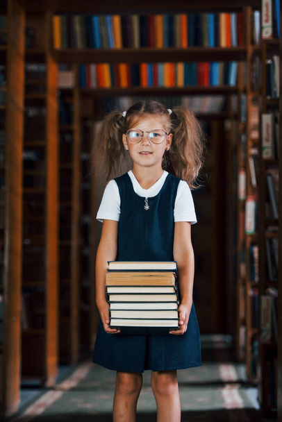 Cute little girl in glasses stands in the library full of books. Conception of education. - Fotó, kép