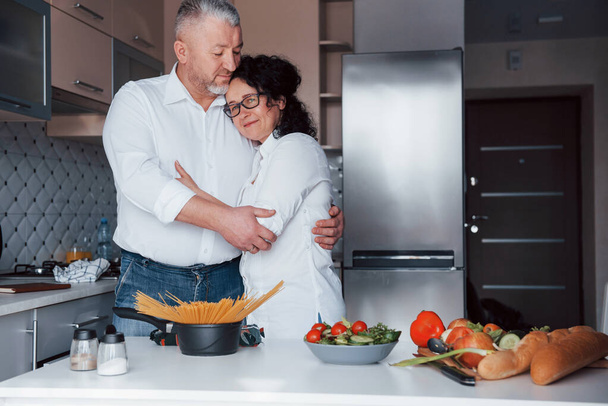 Modern interior. Man and his wife in white shirt preparing food on the kitchen using vegetables. - Photo, image