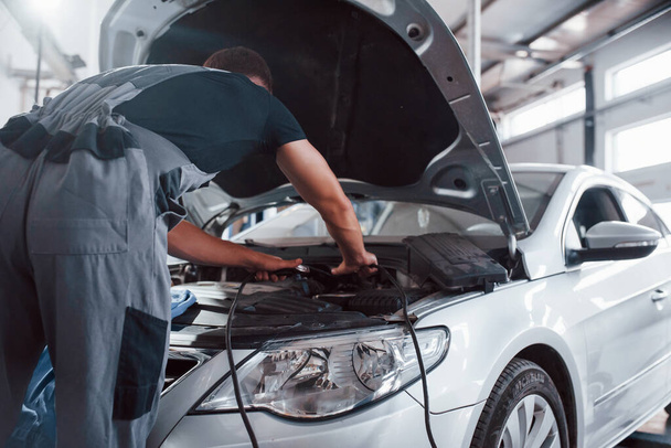 Opened hood. Man in grey uniform repairs white automobile indoors. - Φωτογραφία, εικόνα