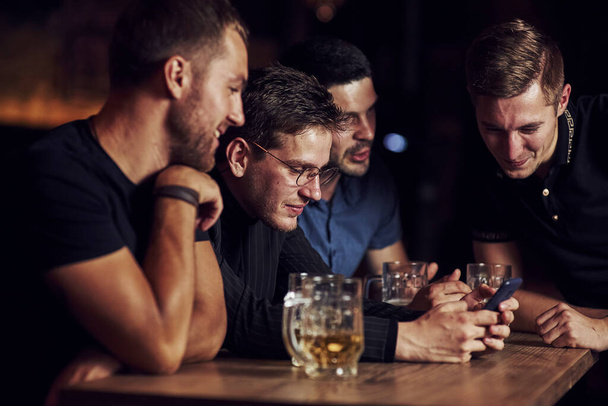 Funny content on smartphone. Three friends resting in the pub with beer in hands. Having conversation. - Fotografie, Obrázek