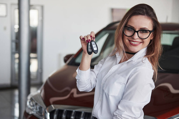 Holds keys in the hand. Woman in eyewear and white shirt stands near the red automobile. - Foto, imagen