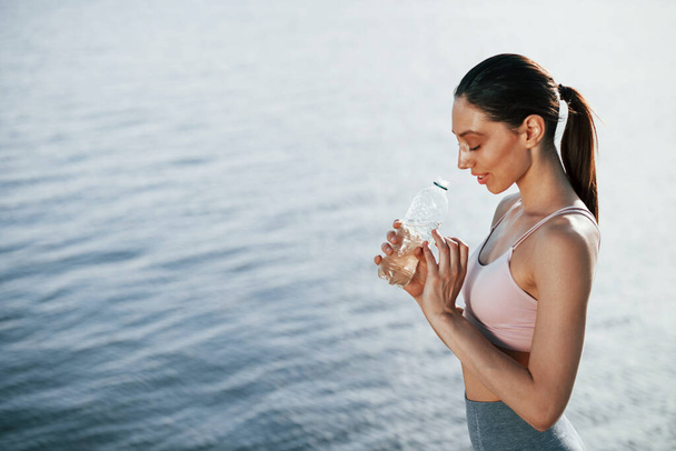 With bottle of water. Girl in sportive clothes stands against the lake. - Foto, imagen