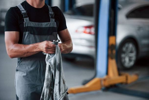 Preparing for the job. Man in black shirt and grey uniform stands in garage after repairing broken car. - 写真・画像