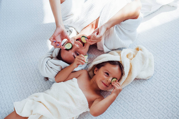 Young positive mother and daughter with towel on head lying on the bed together with cucumber. - Photo, image