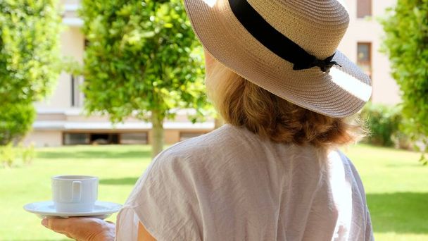 femmes âgées de 50 à 55 ans dans un chapeau de paille tient une tasse de boisson chaude et boit du café, assis le matin sur une terrasse d'été par une journée ensoleillée. Le concept de bonjour, de voyage et d'attente de nouveaux - Photo, image