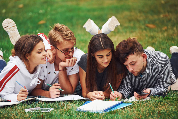 Writing on paper. front view. Group of young students in casual clothes on green grass at daytime. - Foto, Bild