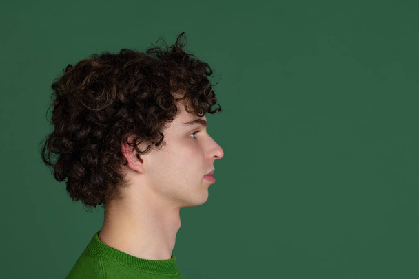 クロップドサイドビューポートレートの若いです男で緑のセーター見ます離れて隔離された上にダークグリーンの背景 - 写真・画像