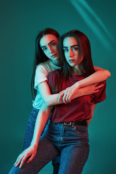 Casual wear. Studio shot indoors with neon light. Photo of two beautiful twins. - Zdjęcie, obraz