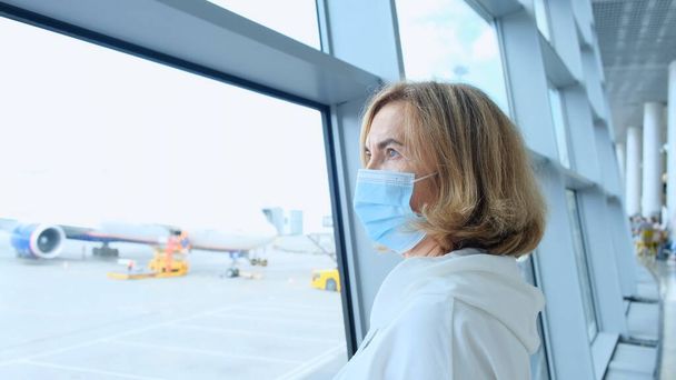 adulto Feminino usando máscara protetora está de pé na janela do terminal do aeroporto aguardando a partida de um voo devido a restrições de viagem devido à pandemia de coronavírus, um idoso de 50 anos - Foto, Imagem