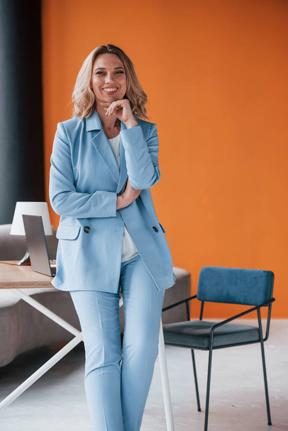 Sincere smile. Businesswoman with curly blonde hair indoors in room with orange colored wall and wooden table. - Photo, Image