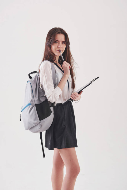 Young female student in skirt and backpack with notepad in hands stands against white background. - Valokuva, kuva