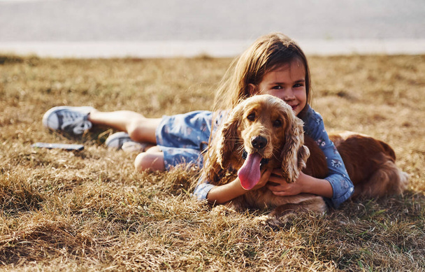 Close up portrait. Lying down on grass. Cute little girl have a walk with her dog outdoors at sunny day. - Foto, Imagem