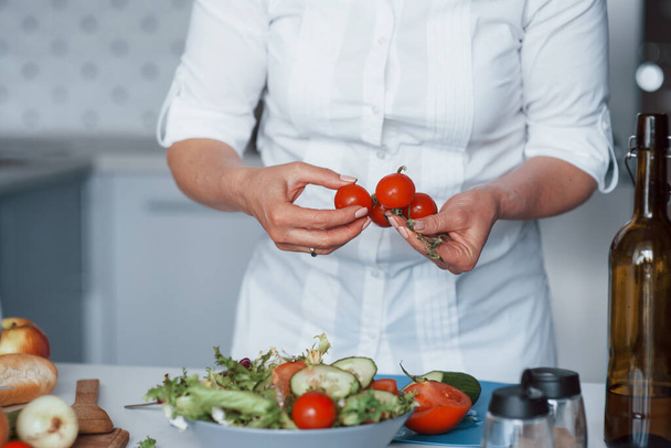 Holding little tomatoes. Woman in white shirt preparing food on the kitchen using vegetables. - Valokuva, kuva