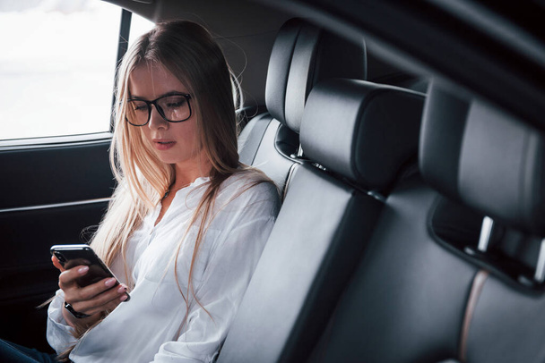 Watching videos on the mobile phone. Smart businesswoman sits at backseat of the luxury car with black interior. - Foto, imagen