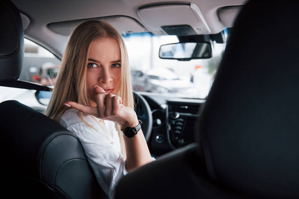 Turned back to the cameramen at backseat. Beautiful blonde girl sitting in the new car with modern black interior. - Photo, Image