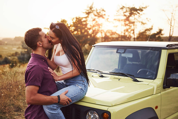 Una pareja encantadora besándose al aire libre. La chica se sienta en un jeep verde. Hermosa noche soleada. - Foto, Imagen