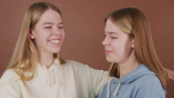 Portrét šťastné mladé dvojče sestry s úsměvem a objímání při pózování pro kameru proti hnědému pozadí - Záběry, video