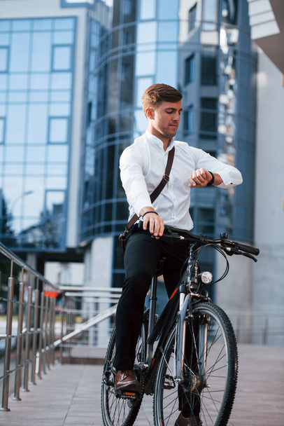 Looks at watch. Businessman in formal clothes with black bicycle is in the city. - Foto, immagini