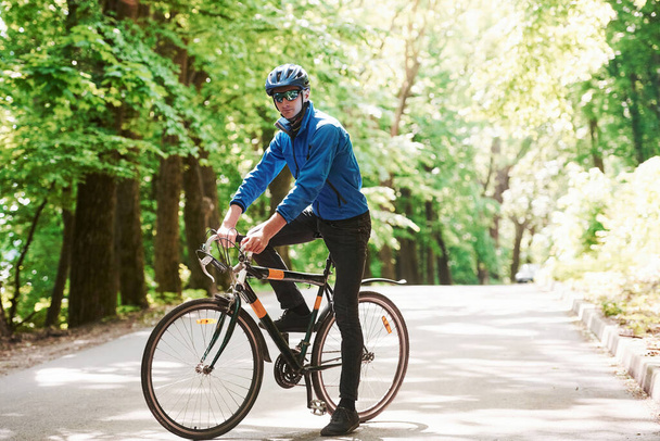 Posing for a camera. Cyclist on a bike is on the asphalt road in the forest at sunny day. - Fotó, kép