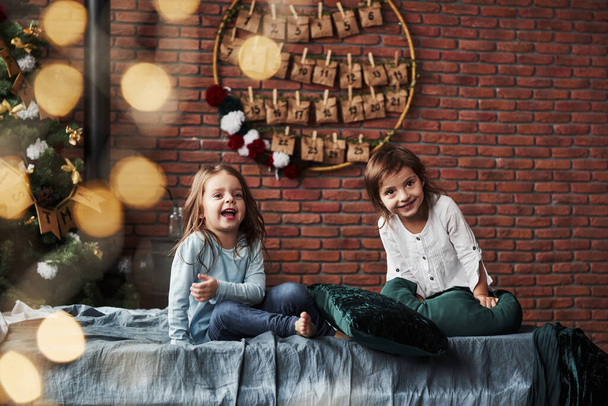 Looking to the side and smiling. Little girls having fun on the bed with holiday interior at the background. - Foto, afbeelding