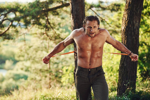 Doing workout. Handsome shirtless man with muscular body type is in the forest at daytime. - Valokuva, kuva