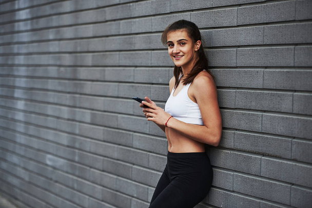 Cheerful mood. With smartphone in hands. Young sportive brunette with slim body shape against brick wall in the city at daytime. - Photo, image