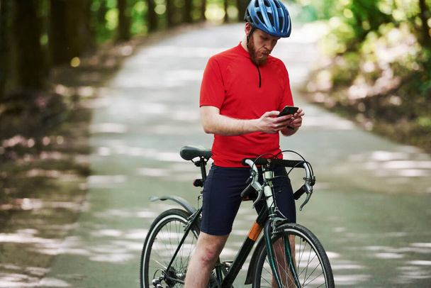 Modern device. Cyclist on a bike is on the asphalt road in the forest at sunny day. - Valokuva, kuva