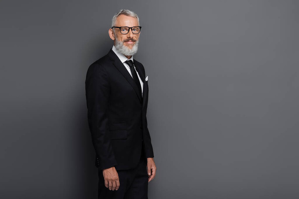 pleased middle aged businessman in glasses and suit looking at camera isolated on grey - 写真・画像