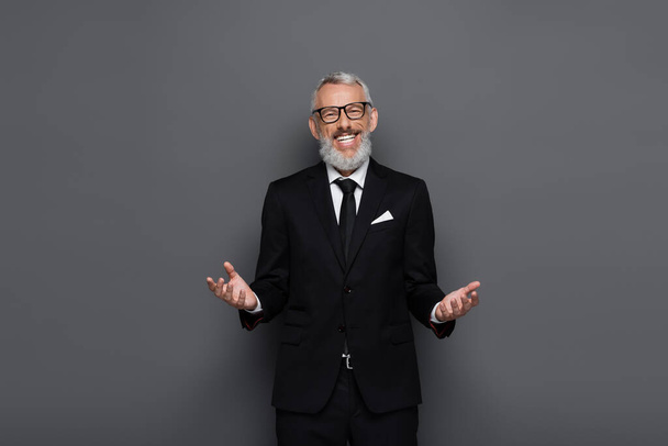 pleased middle aged businessman in glasses and suit looking at camera with welcoming gesture isolated on grey - Foto, immagini