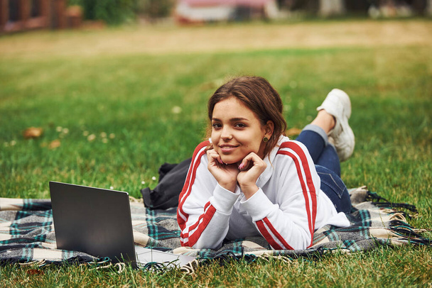 Positive girl in casual clothes near her laptop lying on green grass. - Фото, изображение