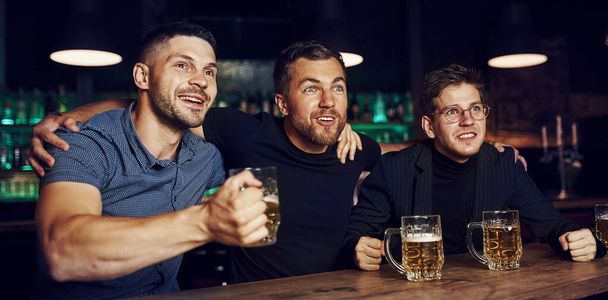 Three sports fans in a bar watching soccer. With beer in hands. - Foto, Bild