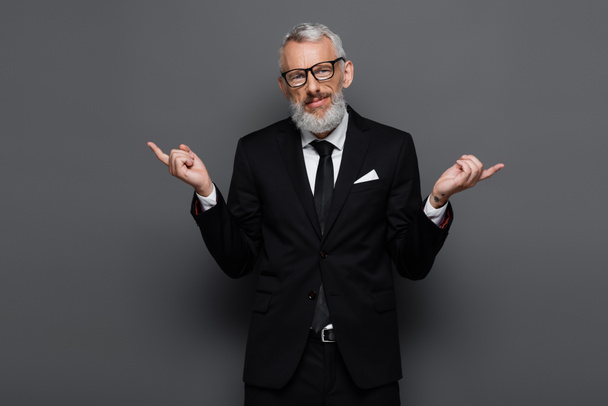 bearded middle aged businessman in glasses pointing with fingers aside on grey - Fotó, kép