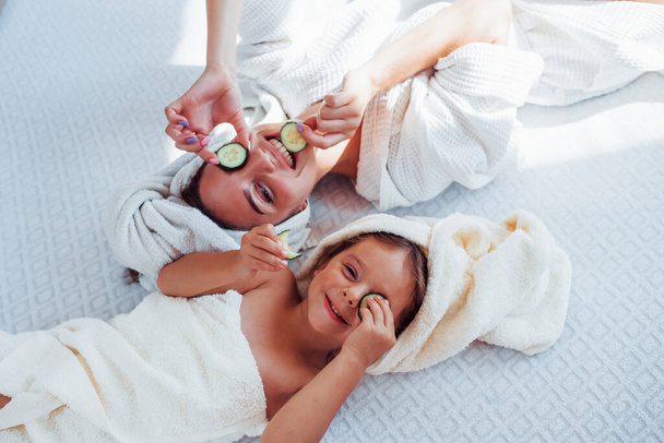 Young positive mother and daughter with towel on head lying on the bed together with cucumber. - Foto, immagini