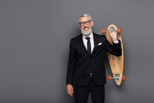 joyful middle aged businessman in suit and glasses holding longboard on grey  - Foto, Imagen