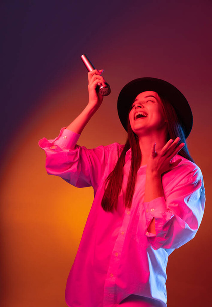 One young beautiful girl wearing black hat using phone isolated on dark studio background in neon light. Concept of emotions, fashion - Фото, зображення