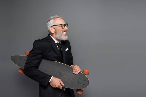 joyful middle aged businessman in suit and glasses holding longboard on grey  - Fotografie, Obrázek