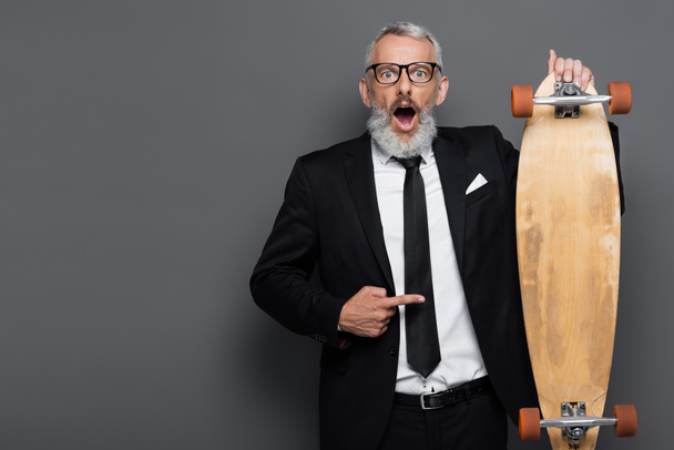 shocked middle aged businessman in suit and glasses pointing at longboard on grey  - Foto, Imagem