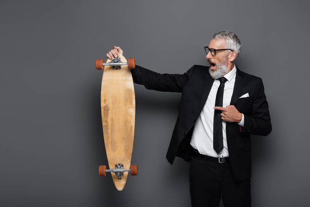 shocked and mature businessman in suit and glasses pointing at longboard on grey  - Fotó, kép