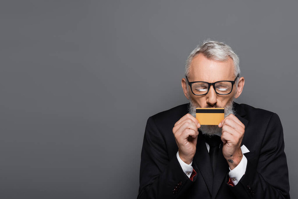 mature businessman in glasses kissing credit card isolated on grey  - Zdjęcie, obraz