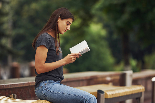 Young brunette sits on the bench and reads book. Trees at background. - Фото, изображение